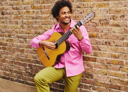 Plínio Fernandes bespeelt zijn gitaar terwijl hij leunt tegen een bakstenen muur. Hij draagt een kleurrijke outfit en kijkt lachend naar opzij.