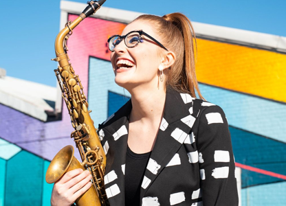 Jess Gillam houdt haar saxofoon vast en kijkt lachen naar boven. Ze draagt een bril, haar haren zitten in een hoge staart en ze draagt een jasje met zwart/witte print. Ze staat voor een kleurrijk gebouw.
